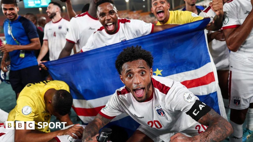 Cape Verde footballers celebrate a victory, with captain Ryan Mendes, wearing a white shirt with red trim, crouching down and extending his arms in front of a national flag and pulling a wide-eyed face of celebration with his mouth wide open