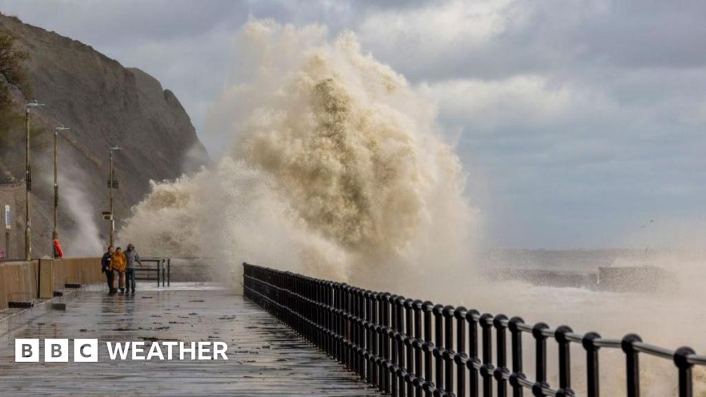 Large waves crashing over a sea barrier with a few people walking along the promenade