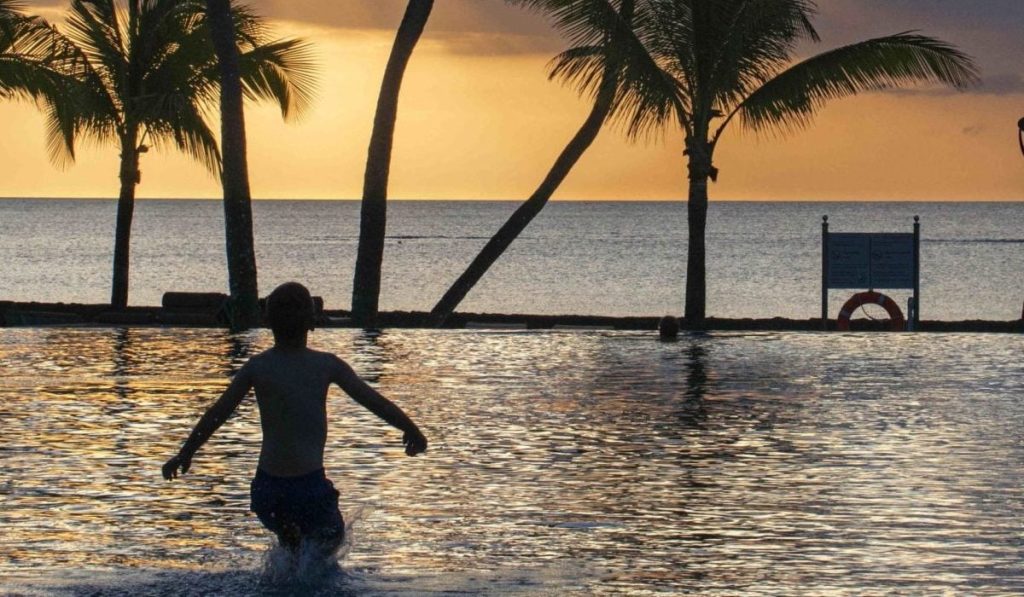 Mauritius, o destinație populară de vacanță. Apus de soare, cu palmieri in fundal și un băiat care intră în apă.